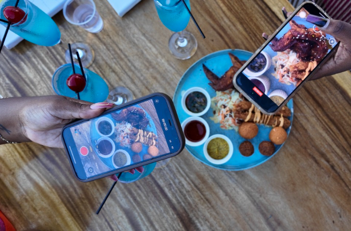 Two people photograph a platter of snacks and sauces, with blue cocktails on the table.