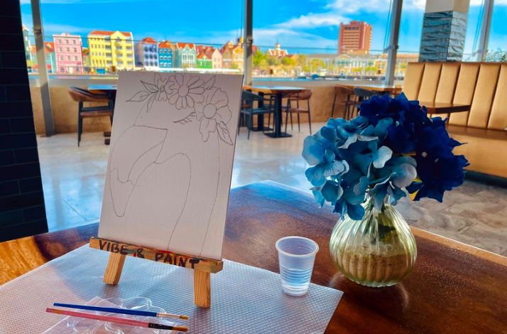 A canvas with a flamingo sketch, paintbrushes, and blue flowers set against Curacao’s colorful waterfront skyline.