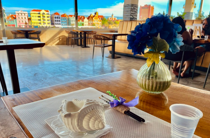 A table set with a ceramic shell, paintbrushes, and a blue flower vase, facing Curacao’s colorful skyline.