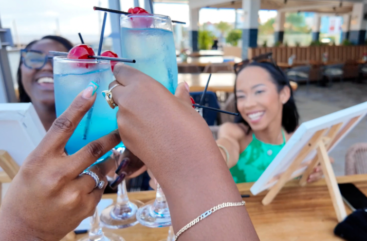 Blue cocktails with cherries are raised in a toast beside painting easels at a waterfront venue.