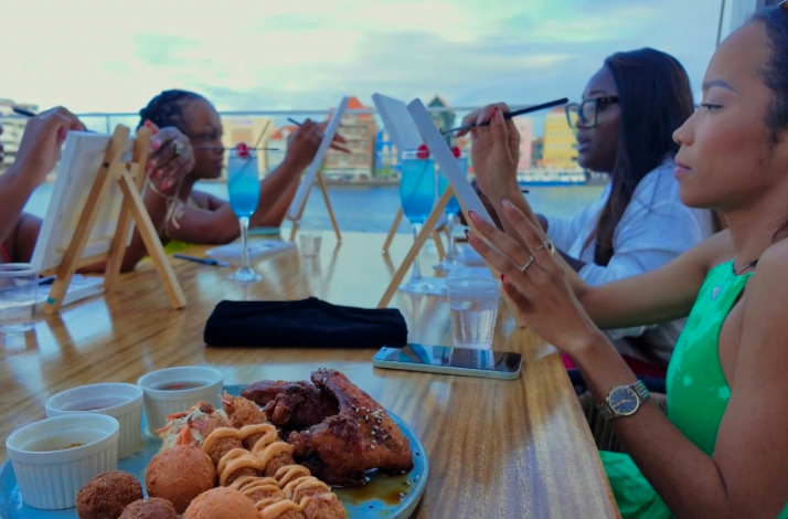 Guests paint at easels with blue cocktails and snacks, with Curacao’s vibrant buildings in the background.