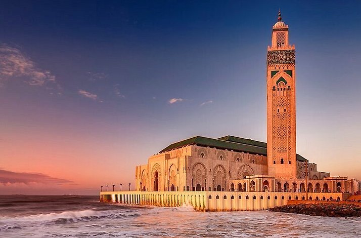 Hassan II Mosque stands majestically by the ocean, bathed in warm sunset light and waves below.