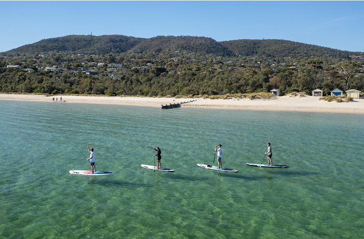 Four people paddleboarding on calm, clear water near a sandy beach and forested hills at the Mornington Peninsula, Australia.