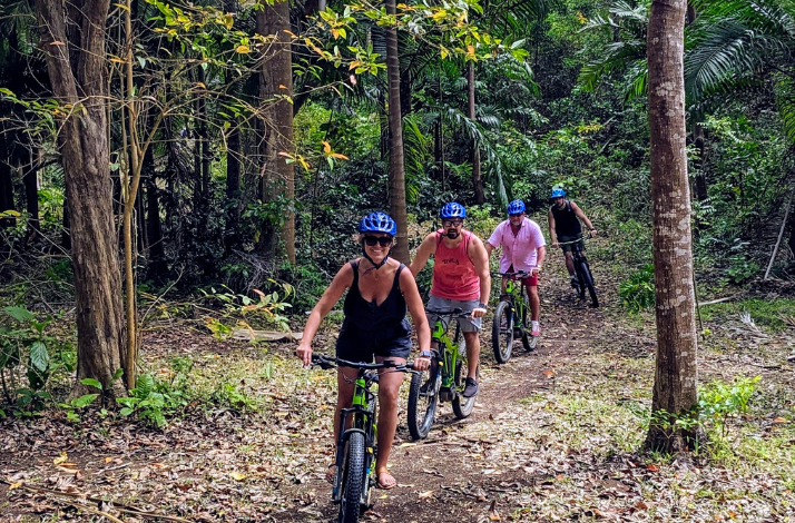 A group of four people rides their bicycles along a trail in a lush forest of Barbados, with each person wearing a helmet