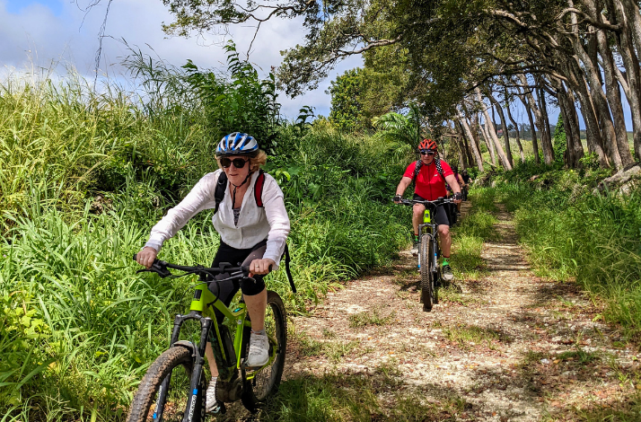 Two cyclists ride mountain bikes along a dirt path through a lush, green, sunlit forest in Barbados