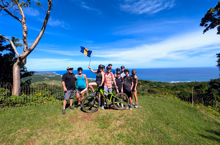 A group of people posing with a bicycle on a hilltop overlooking a beautiful coastal view, whilst holding a Barbados flag