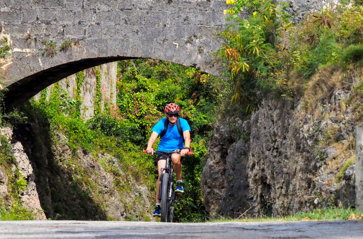 A cyclist, wearing a helmet, rides his mountain bike  under a stone arch bridge in Barbados