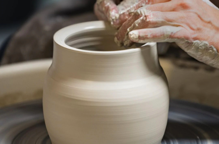 A close-up shot of a person's hands shaping a wet clay pot on a spinning pottery wheel.