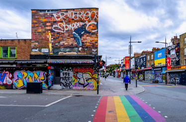 A vibrant city street with colorful murals on building walls and a rainbow-painted crosswalk  featuring a portrait of local guide Tom