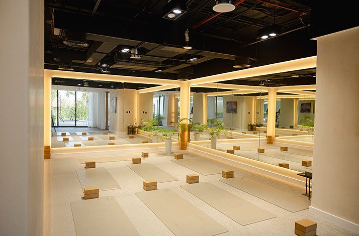 Spacious yoga studio interior with rows of mats and cork blocks, warm LED lighting, mirrored walls, and tropical plant accents.