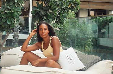 Adrienne Everett seated on an outdoor daybed in a yellow swimsuit, beside an 'h. Studio' logo pillow, surrounded by tropical greenery.