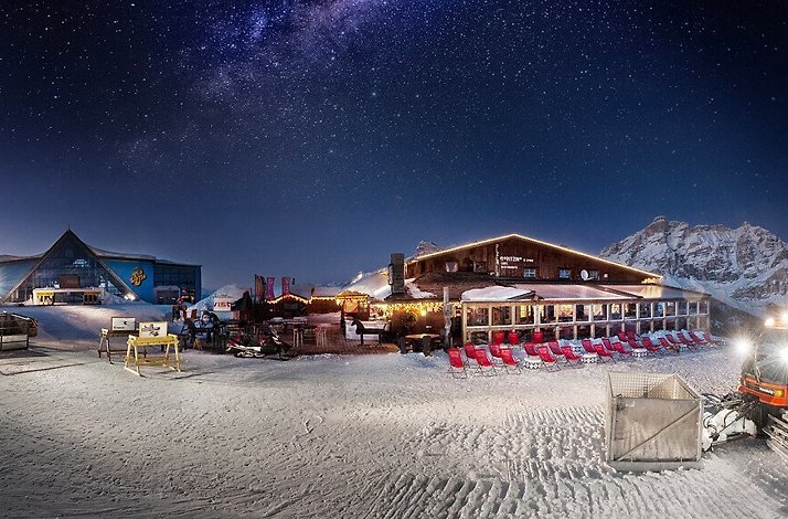 Panoramic view depicts a nighttime scene at Club Moritzino, showcasing buildings, equipment, and the starry night sky. 