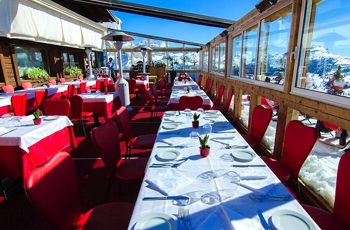 Club Moritzino restaurant patio with red tables and seats set for dining, with view of the mountains through its window glass. 