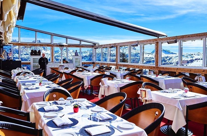 Club Moritzino restaurant patio with wooden tables and seats set for dining, with view of mountains through its window glass.