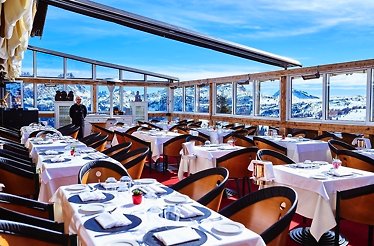 Club Moritzino restaurant patio with wooden tables and seats set for dining, with view of mountains through its window glass.