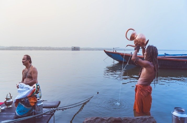 Two men stand at the water's edge, with a boat visible in the background under a clear sky.