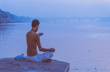 A man sits on the riverbank, looking at his phone while enjoying the view of the flowing water.