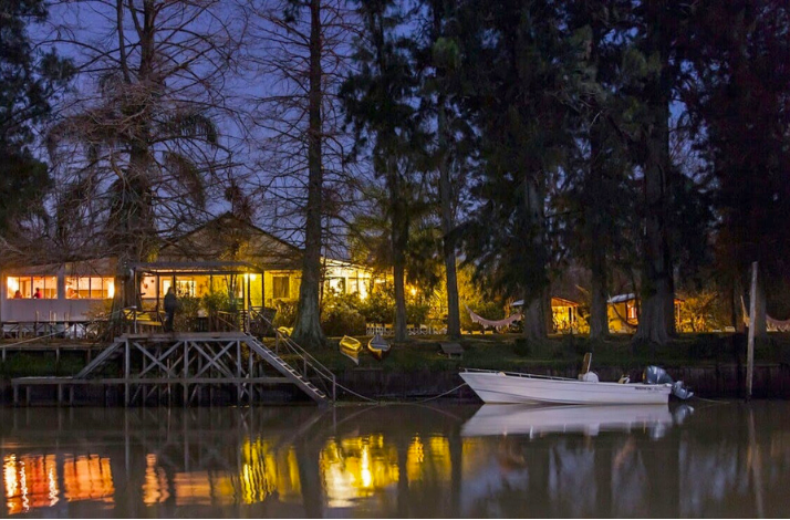 Riverside lodge warmly lit at night, with a boat docked on the calm water.