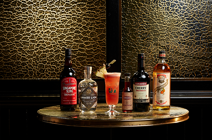 Singapore Sling cocktail on a round table surrounded by bottles of spirits and mixers.