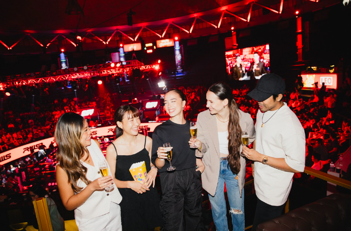 Five young people smiling and enjoying drinks and snacks on a Thai boxing match on the Rajadamnern Stadium in Bangkok Thailand.