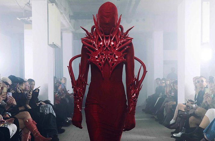 Model walking the runway in Marlon Ferry's all-red sculptural look featuring spiked armor bodice, full hood, and articulated arm pieces.