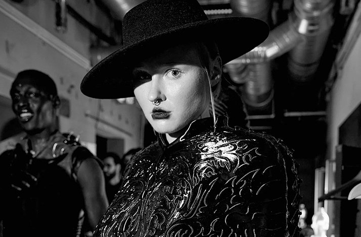 Close-up black-and-white backstage portrait of a model in an ornate embroidered jacket and wide-brim hat.