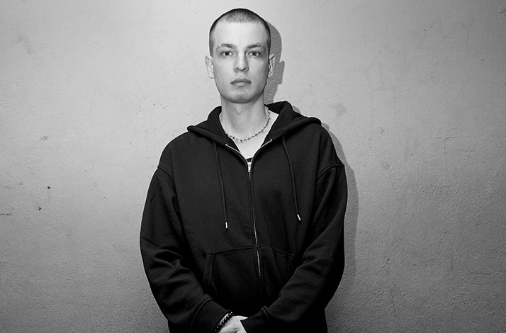 Designer Marlon Ferry standing against a concrete wall, wearing a black zip-up hoodie and chain necklace.
