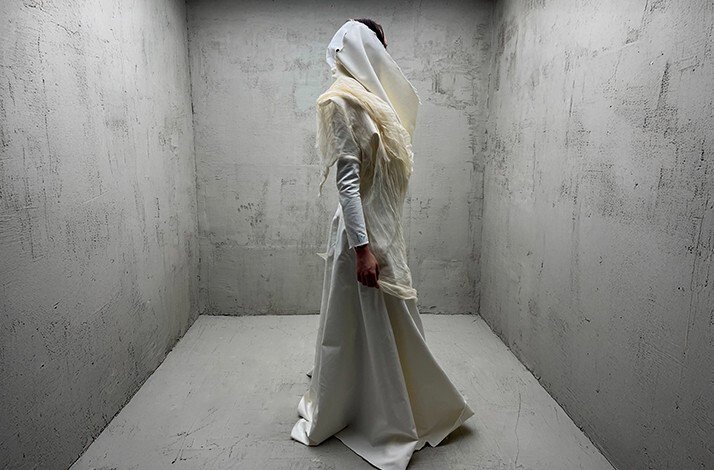 Model wearing HADERLUMP 's floor-length white latex couture gown with draped hood, posed in a concrete-walled studio.