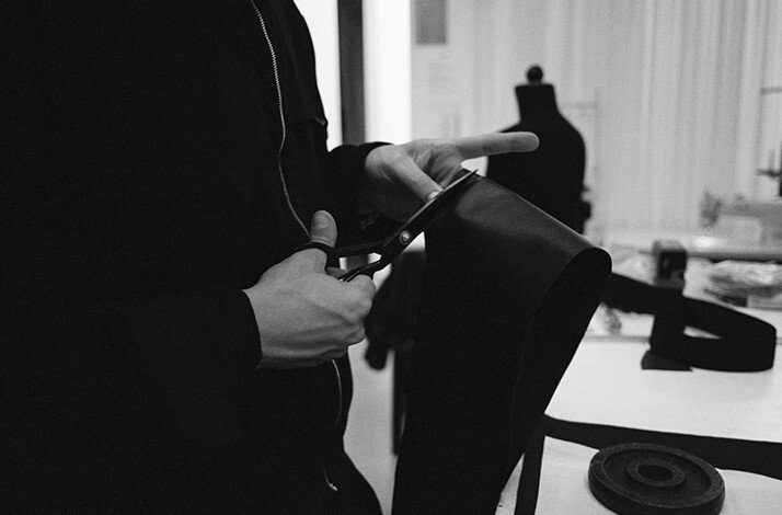Hands cutting dark latex fabric with scissors in HADERLUMP 's atelier, a dress form visible in the blurred background.