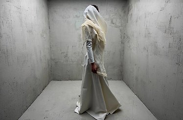 Model wearing Marlon Ferry's floor-length white latex couture gown with draped hood, posed in a concrete-walled studio.