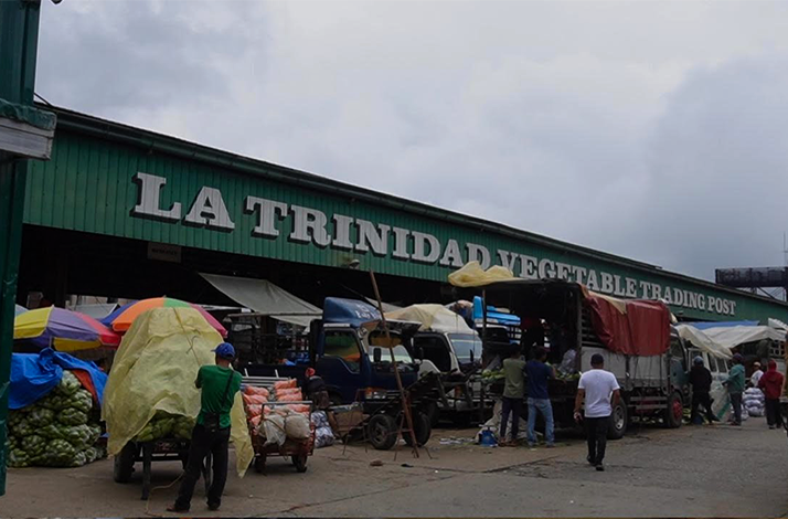 Exterior view of La Trinidad Vegetable Trading Post with trucks, produce sacks, and vendors unloading goods