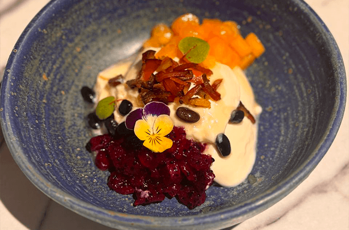 A bowl of creamy dessert topped with fruits, seeds, and an edible flower.