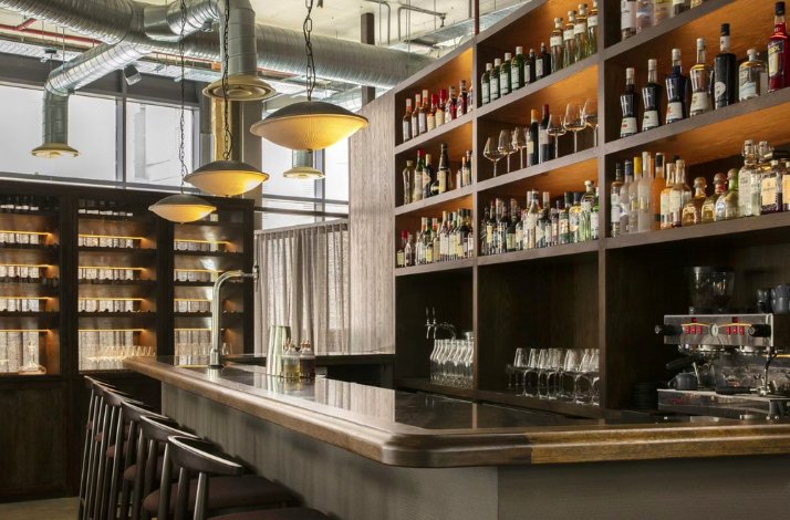 A modern bar interior with a polished counter, high wooden stools, and shelves lined with various bottles of liquor and glassware. Overhead, industrial-style lighting and exposed ductwork add to the contemporary atmosphere.