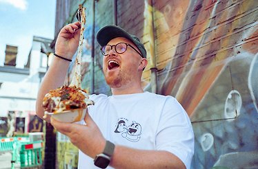 A person in a white t-shirt and cap holds up a bowl of loaded fries with melted cheese stretching upwards, standing in front of a colorful graffiti-covered wall in an outdoor urban setting.