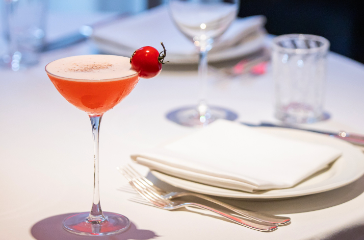 A glass of cocktail with a cherry tomato served next to a white plate with a white napkin for the guest.