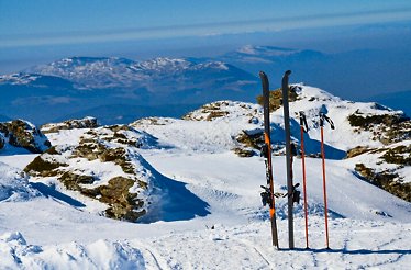 Skis upright in the snow on a mountain, overlooking a scenic view of mountain peaks.