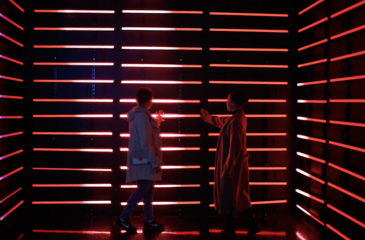 Two people facing each other with hands raised beside walls of red neon strip lights in a darkened room.