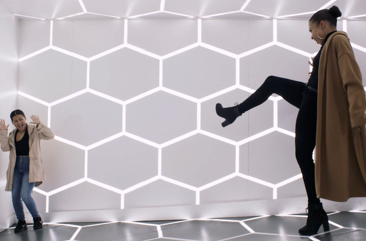 Two visitors laughing and posing beside a floor-to-ceiling illuminated hexagonal light wall.