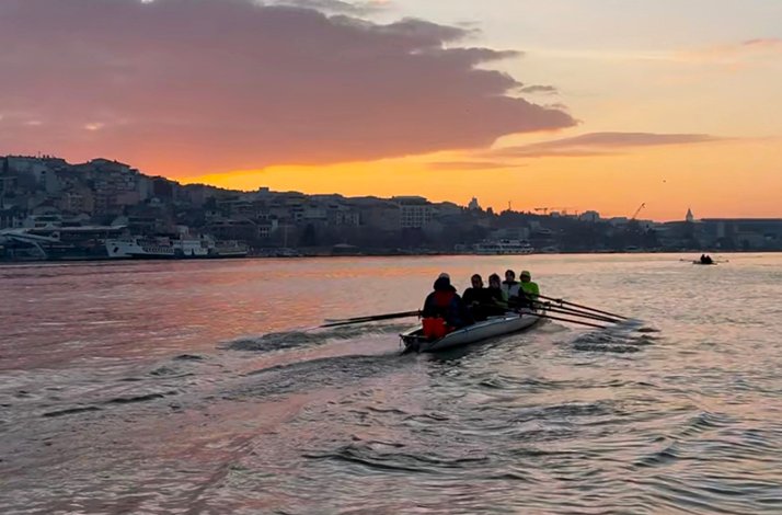 Boat of rowers moving along a river at sunrise near a city shoreline.