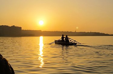 Rowers silhouetted on water at sunset with city skyline in the background.