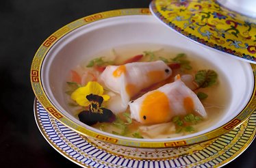 Two handcrafted lobster dumplings shaped like koi fish swimming in a clear supreme broth with edible flowers.