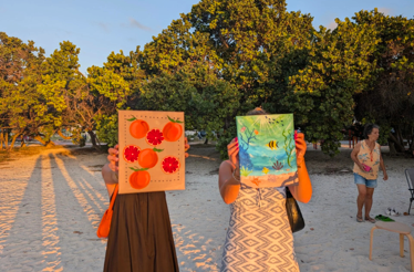 Two people hold up their finished beach‑painted canvases at sunset.