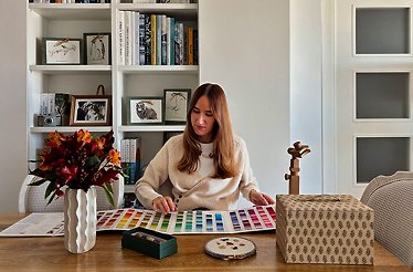 Person selecting thread colors at a table with embroidery supplies, books, and flowers.