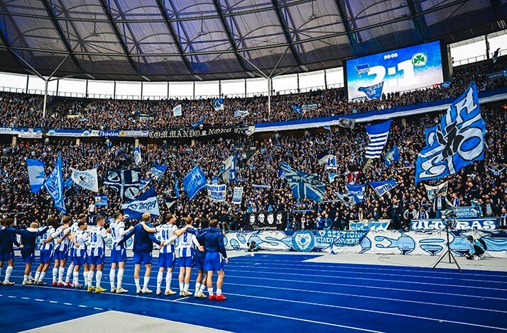 Hertha BSC players in blue-and-white kits stand in a line on the pitch, applauding a packed stadium filled with blue and white flags. The scoreboard reads 2:1.