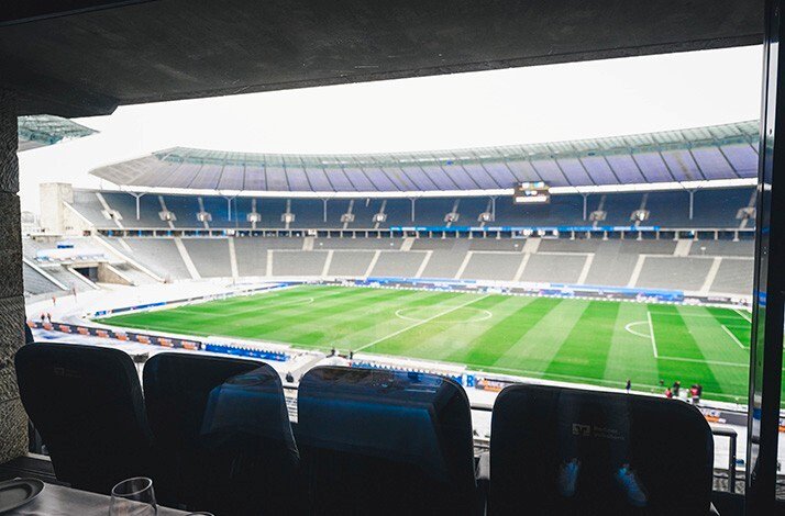 View from a VIP lounge across the empty stadium to the green pitch, framed by dark leather seats and a glass in the foreground.