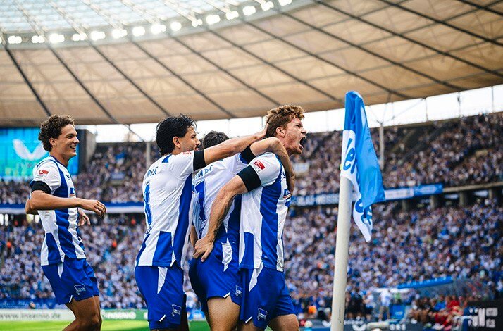 Hertha BSC players in blue-and-white kits celebrate closely together, one shouting with joy, beside a blue corner flag and a cheering crowd.