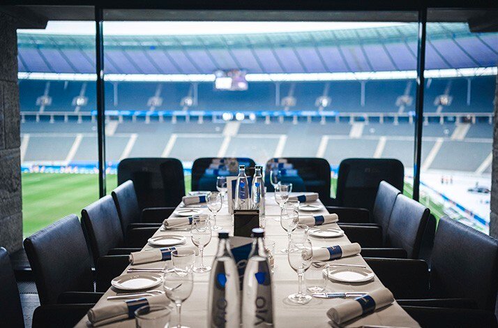 A long dining table set with white plates, folded blue napkins, water bottles, and glasses, with large windows overlooking the Olympiastadion Berlin pitch.