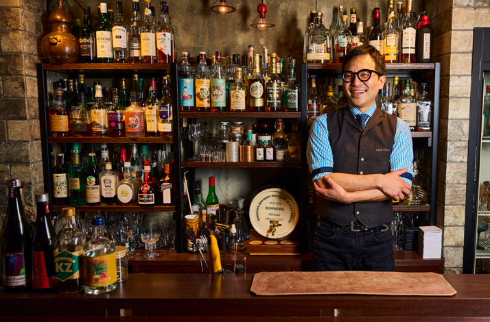 Smiling bartender in a vest and glasses standing with arms crossed in front of shelves stocked with an extensive whiskey and spirits collection.