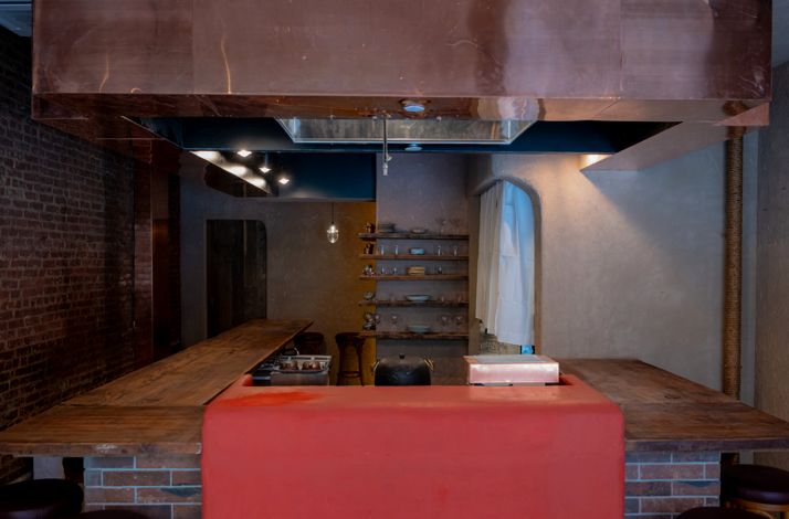 Intimate restaurant interior featuring an open kitchen with a copper hood, exposed brick walls, wooden counters, and a red island station.