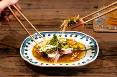 Chopsticks lifting a piece of fish from an elegantly plated Japanese dish in a blue and white ceramic bowl, paired with a cocktail at Odo restaurant.
